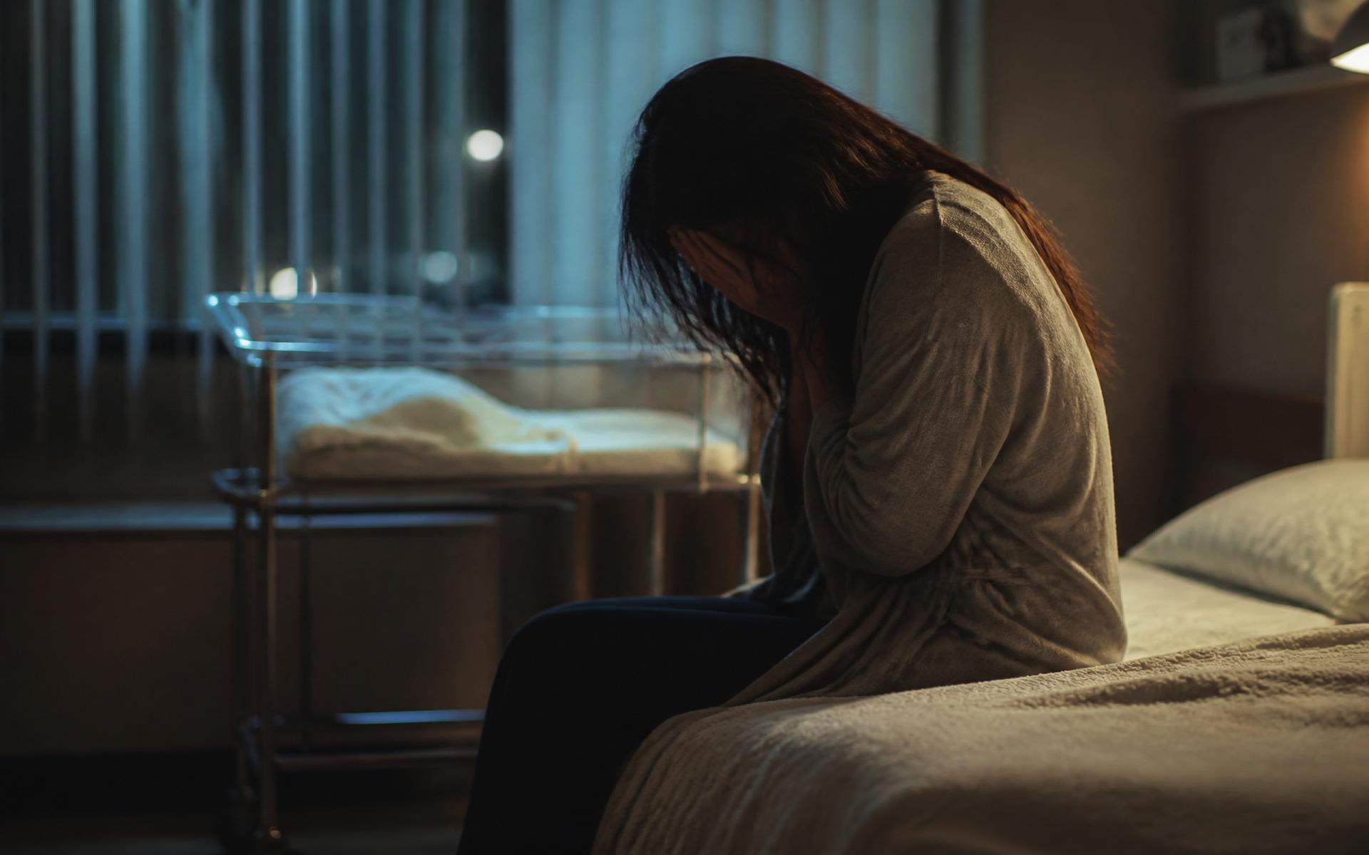 A young mother alone in a dim hospital room, an empty bassinet beside her